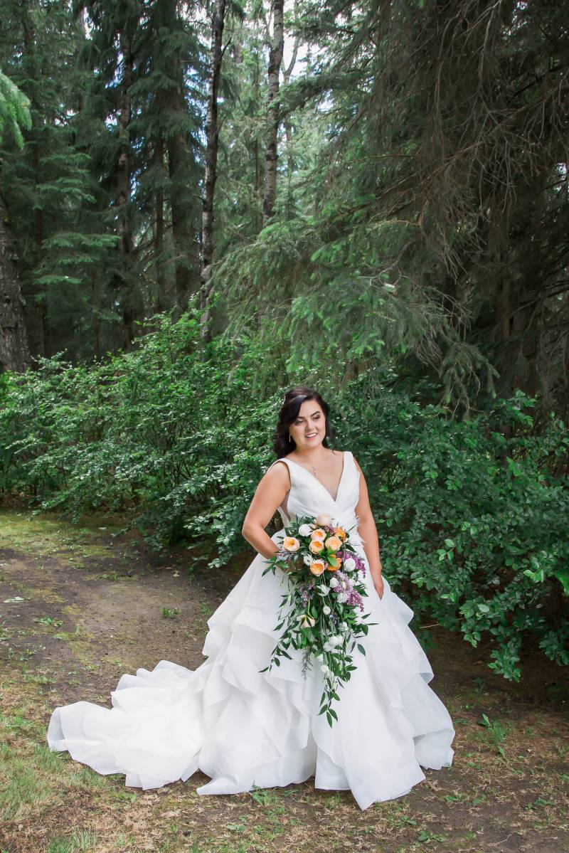 Bride holding yellow purple and white bouquet standing in white dress in front of dark green forest
