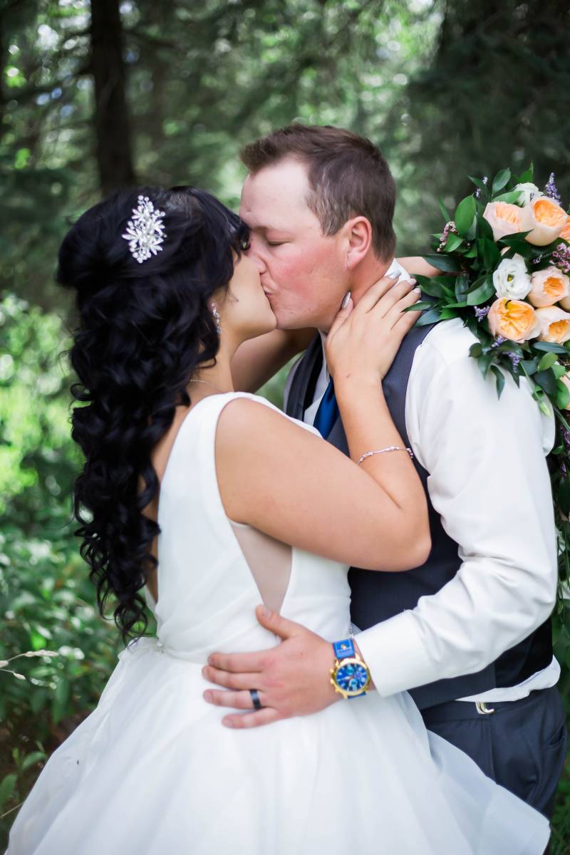 Bride and groom embrace while kissing holding bouquet in front of dark green forest