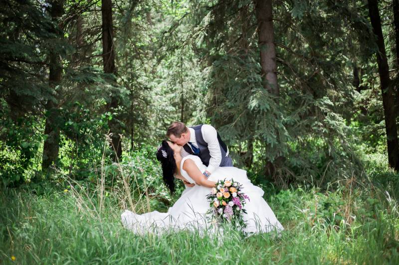 Groom dips bride holding yellow and purple bouquet backing lush green forest