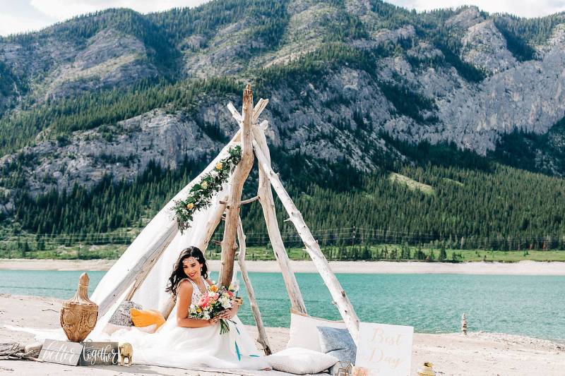 woman sits under drift wood and white veil holding pink and yellow bouquet in front of lake and mountains 