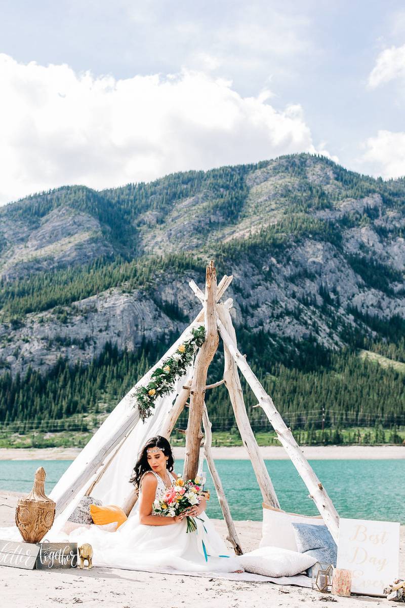 woman sits under drift wood and white veil holding pink and yellow bouquet in front of lake and mountains 
