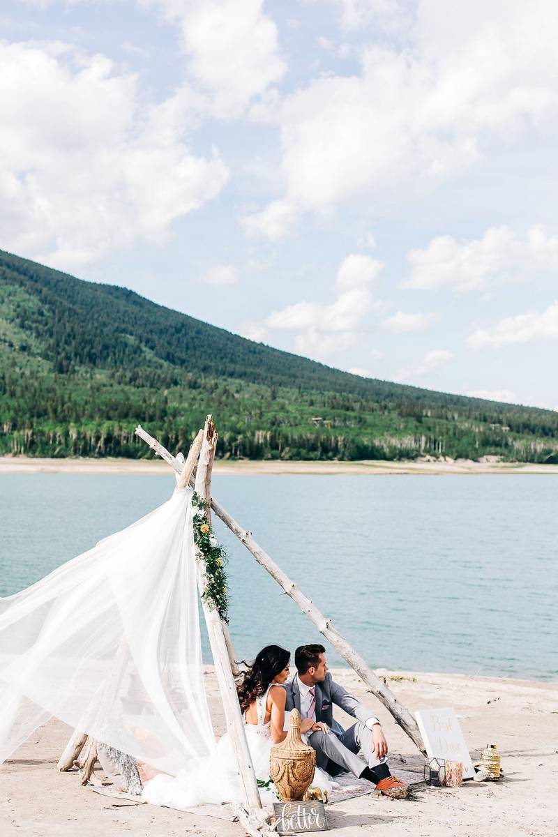 Man and woman sit under driftwood and veil facing lake and mountains 