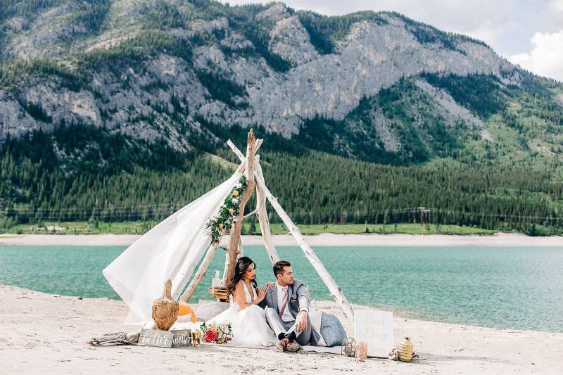Man and woman sit under drift wood and white veil in front of lake and mountains 