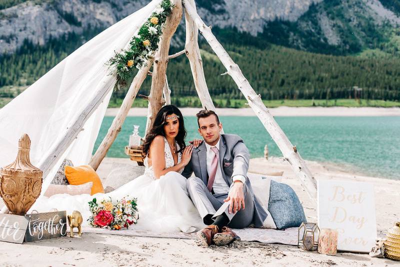 Man and woman sit under drift wood and white veil in front of lake and mountains 
