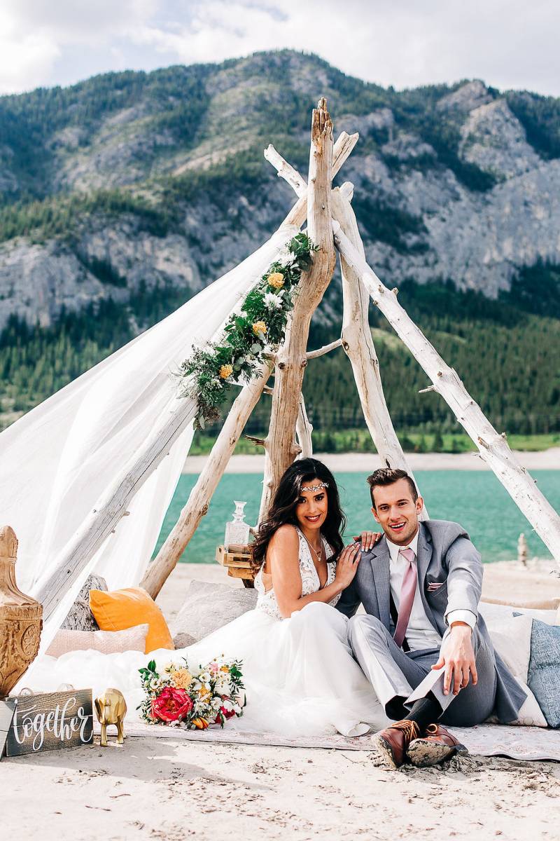 Man and woman sit under drift wood and white veil in front of lake and mountains 