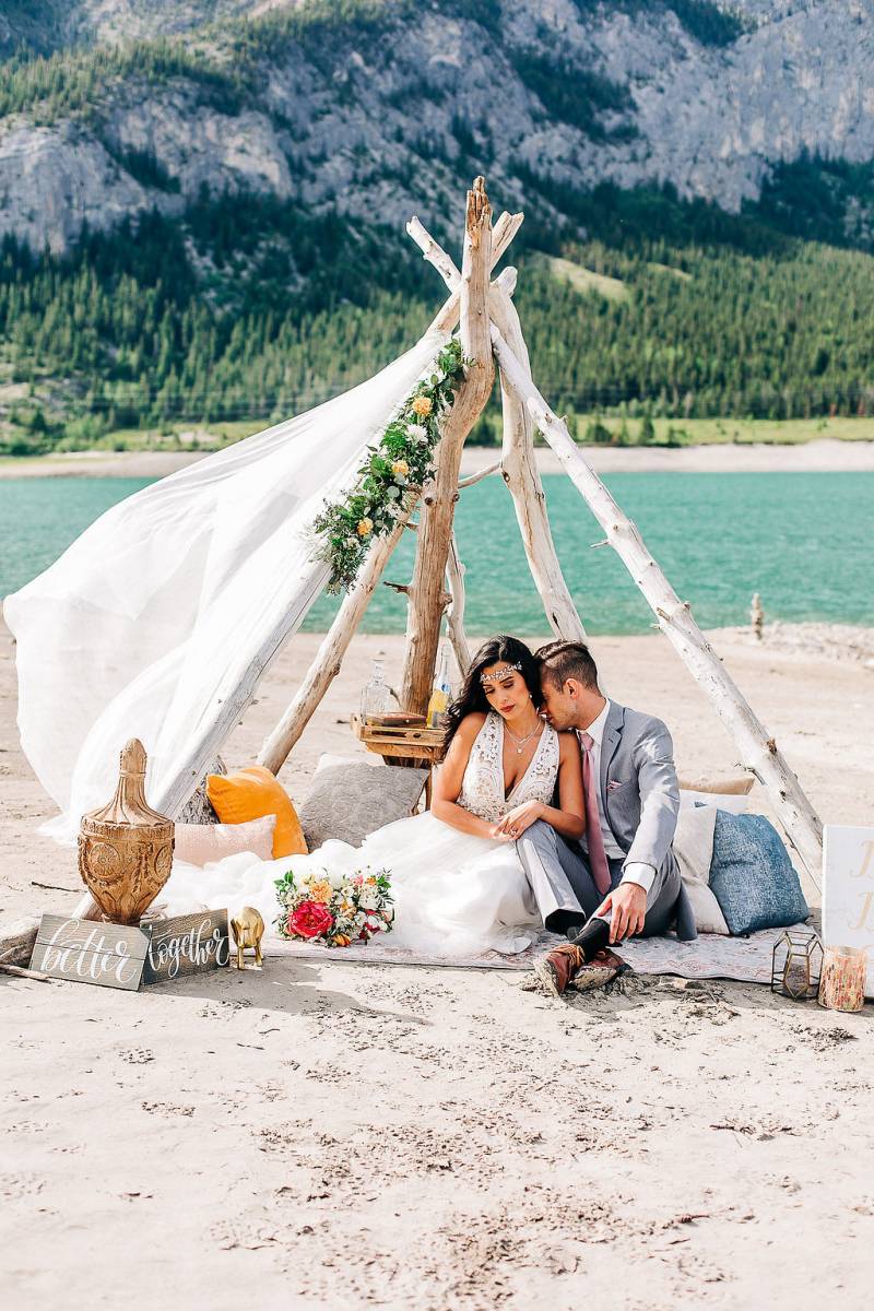 Man and woman sit under drift wood and white veil in front of lake and mountains 
