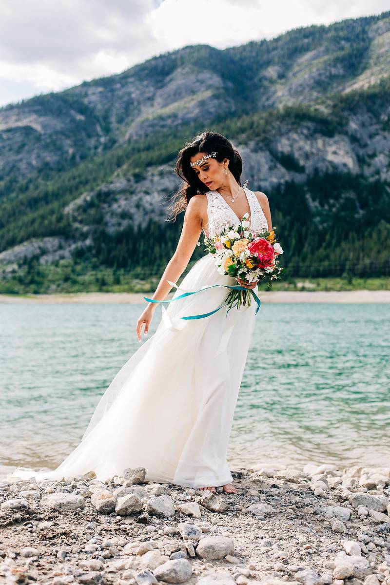 Woman leans over in white dress holding pink and yellow bouquet in front of lake 