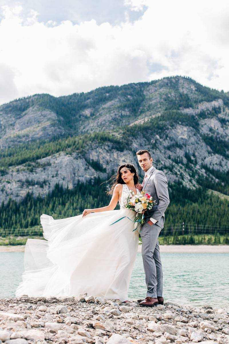 Man and woman stand while white dress blows in wind in front of lake and mountains 