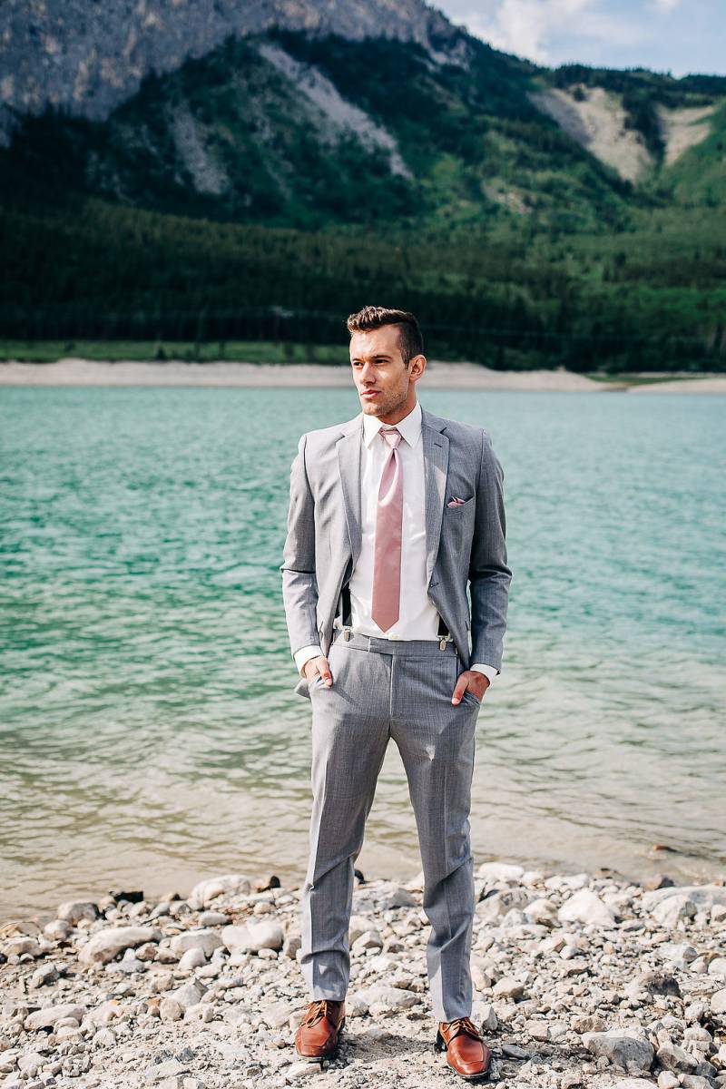 Man stands hands in pockets of grey suit in front of lake and mountains 