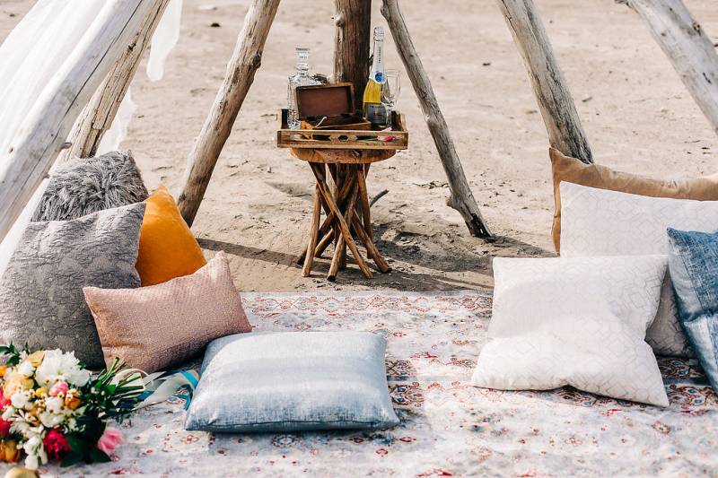 Multicolored pillows on carpet on sand with small table and minibar under driftwood and veil 