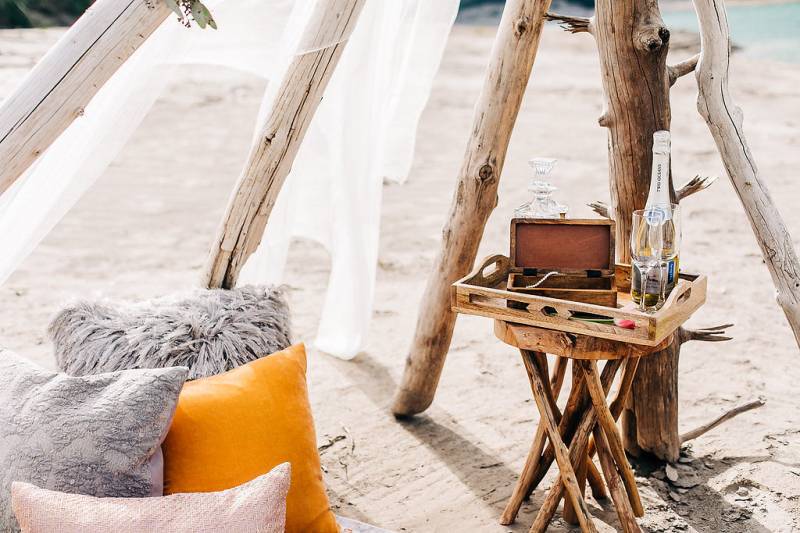Pillows beside Small wooden tray with box and bottle of wine with glasses sitting on small wooden stool 