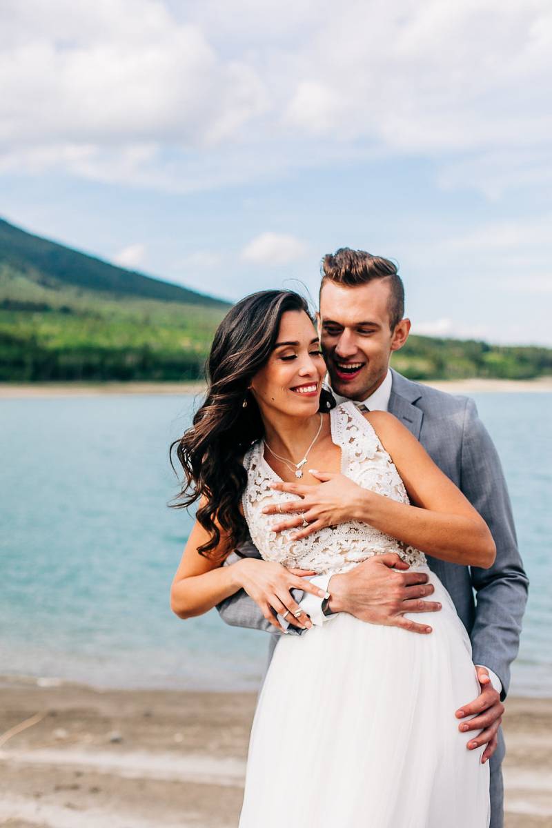 Groom embraces bride from behind smiling holding hip in front of lake 