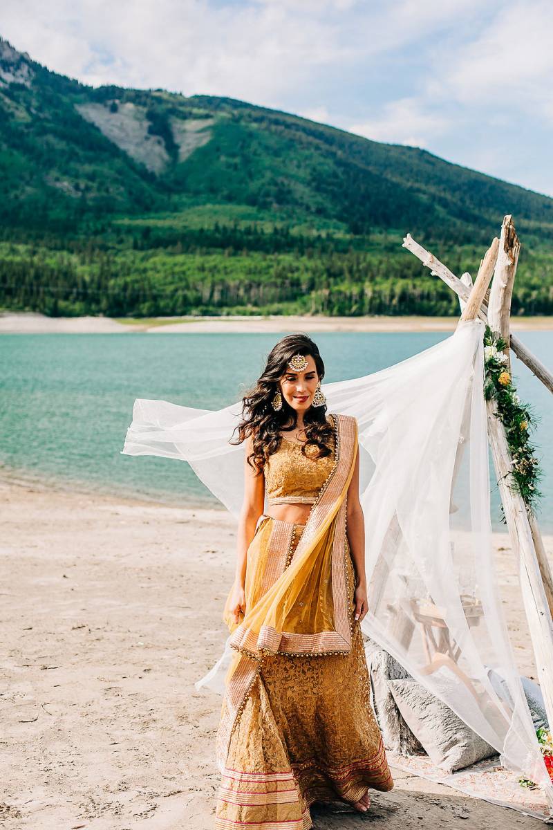 Woman stands in yellow dress in front of white veil tied up with driftwood backing lake 