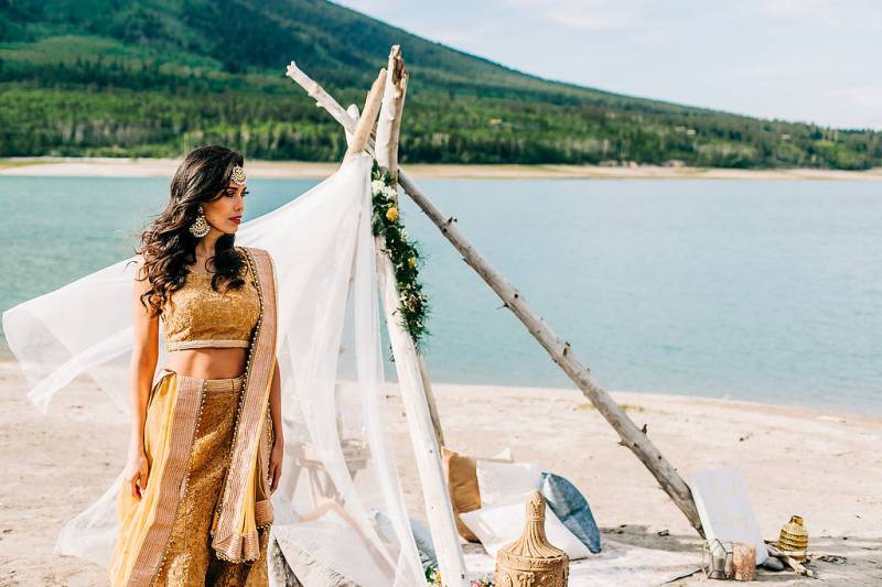 Woman stands in yellow dress in front of white veil tied up with driftwood backing lake 