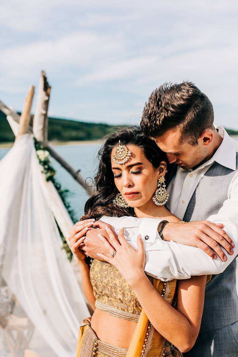 Man embraces woman from behind  beside white veil and driftwood 