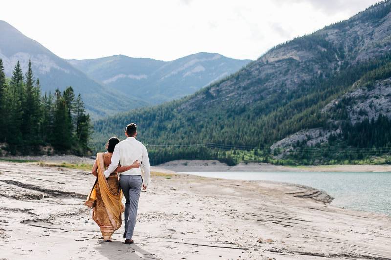 Man and woman walk along edge of water arms around back facing mountains 