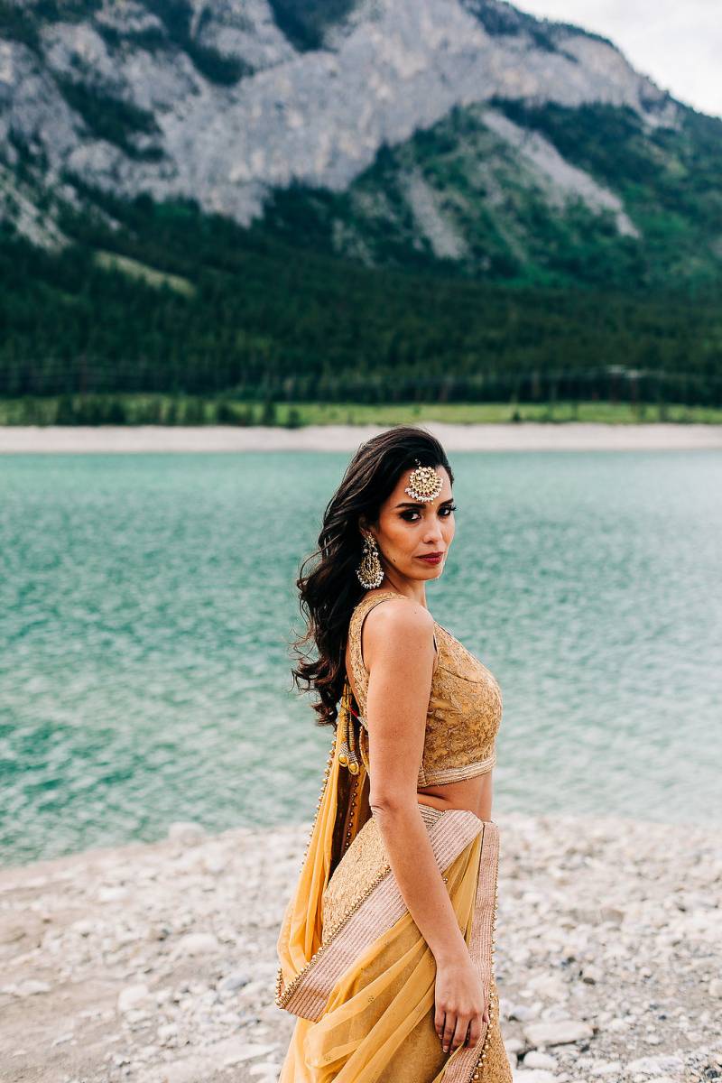 Woman in yellow dress stands on rocky bank overlooking lake and mountains 