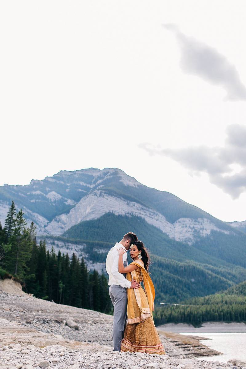 Man in grey pants holds waist of woman in yellow dress on rocky bank backing mountains 