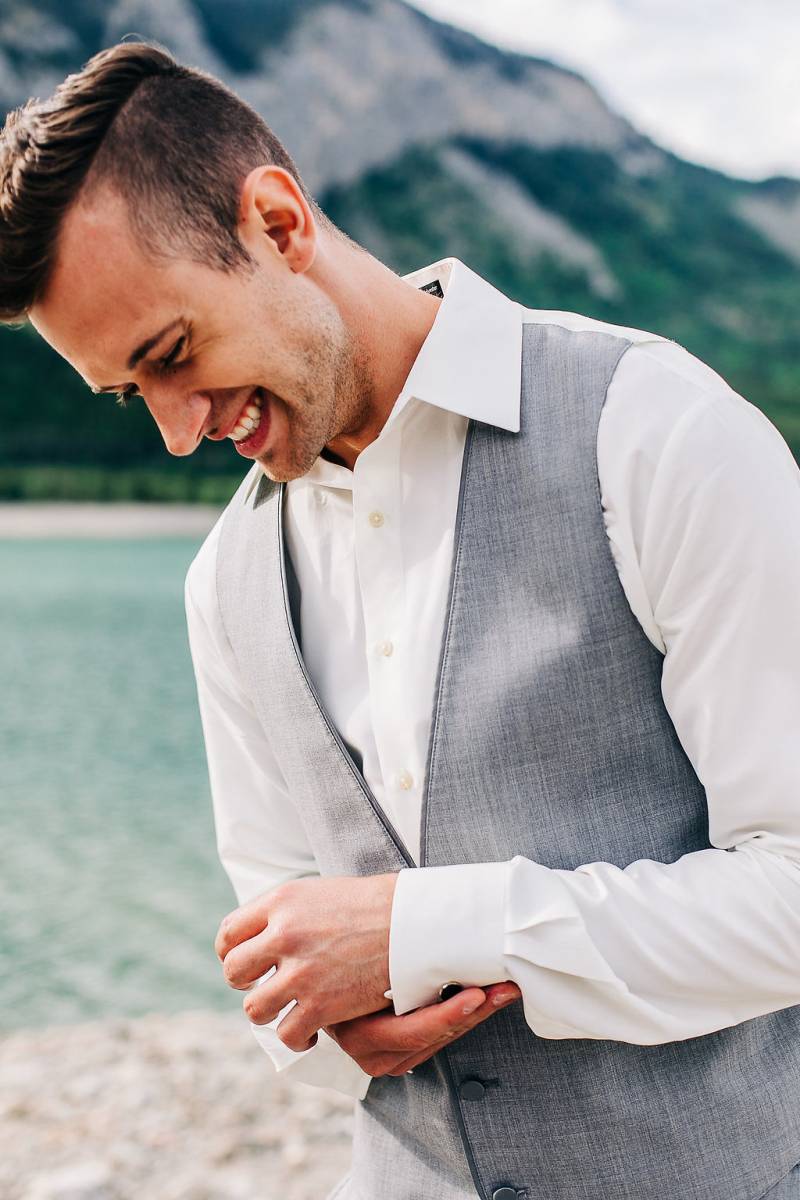 Man in grey vest smiles adjusting shirt cuff looking down 