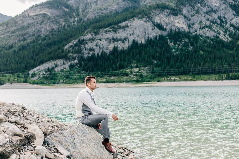 Man in grey suit and shit sits on rock looking towards lake and mountains 