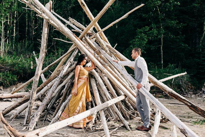 Bride and groom stand amidst large logs stacked into teepee 