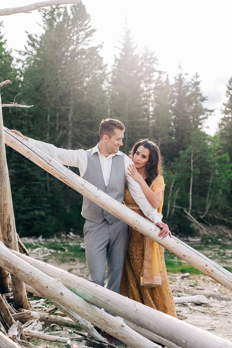 Woman leans against man leaning on long driftwood backing forest 