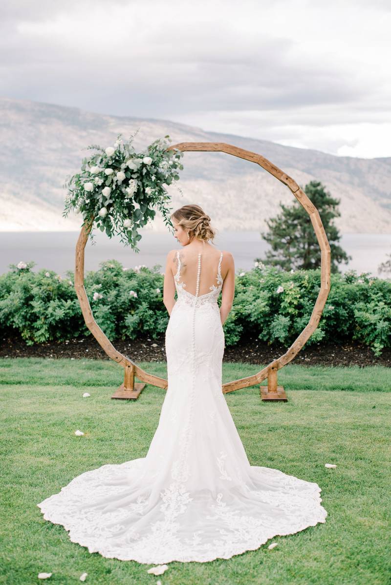 Bride stands facing large wooden wedding arch and large white floral arrangement 