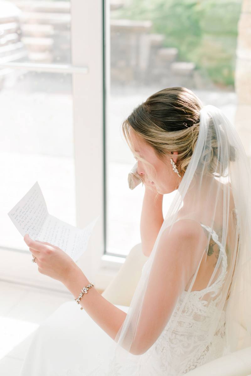 Bride sitting in white dress and veil dabs eyes while reading note