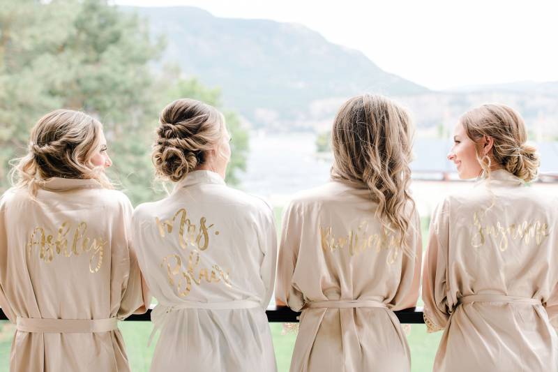 Bride and bridesmaids wearing matching blush robes with gold lettering standing on balcony 