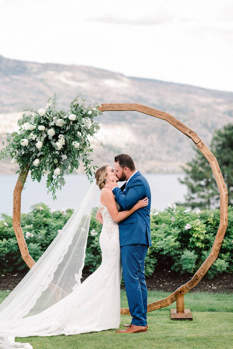 Bride and groom kiss in front of large wooden wedding arch 