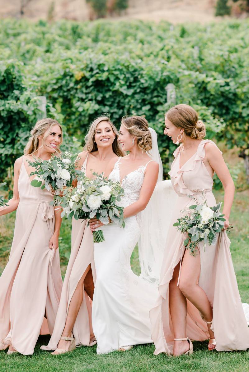 Bride and bridesmaids laughing walking in green field holding white bouquets 