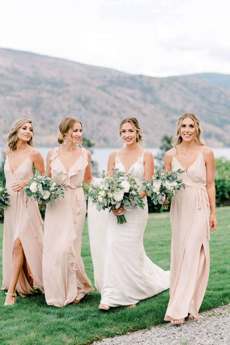 Bride and bridesmaids walking together down grassy field overlooking mountains 