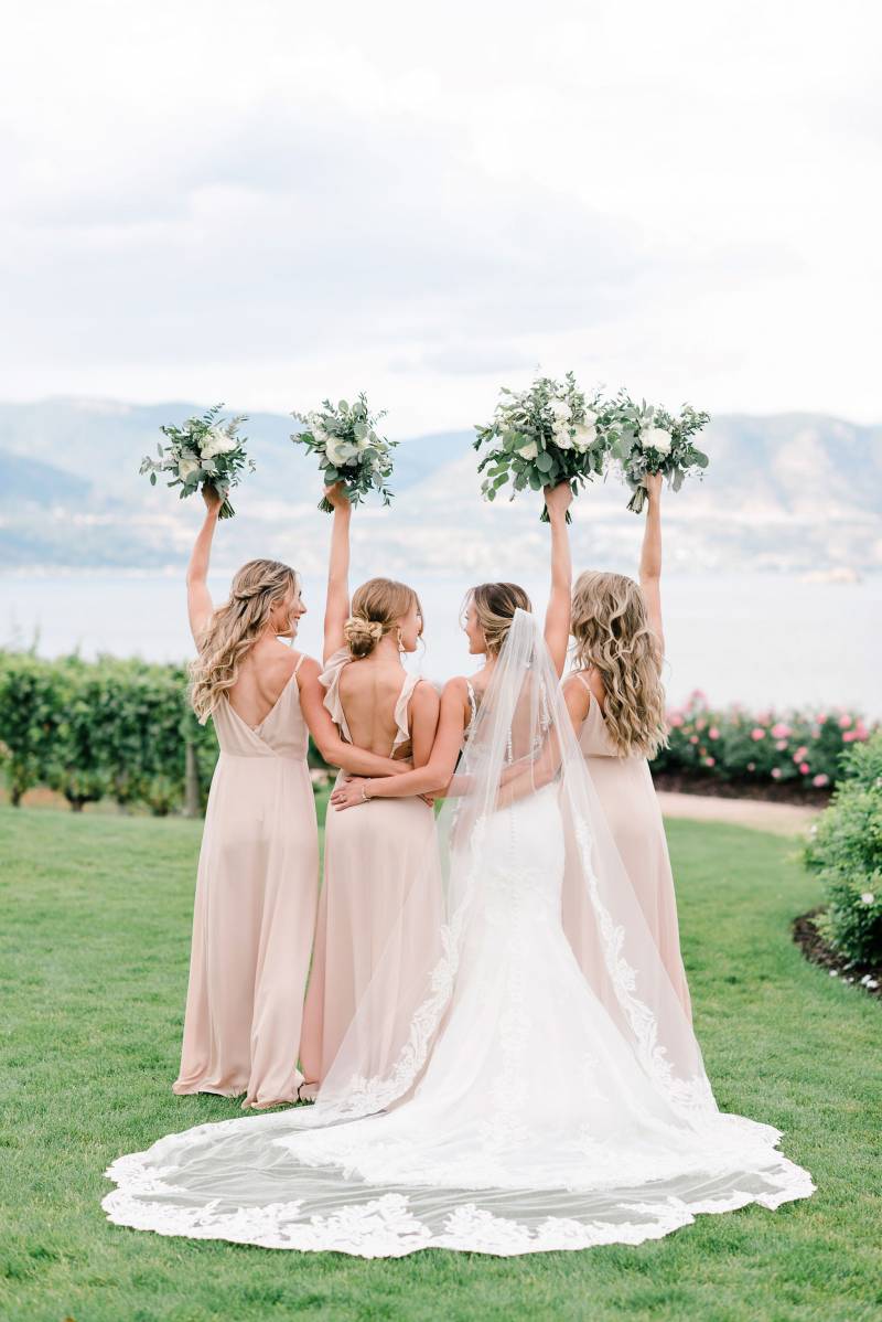 Bride and bridesmaids holding bouquets in air facing mountains and lake 