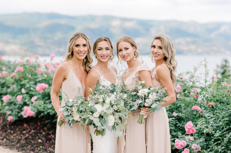 Bride standing with bridesmaids holding white flower bouquets on dirt path in front of pink rose bushes 
