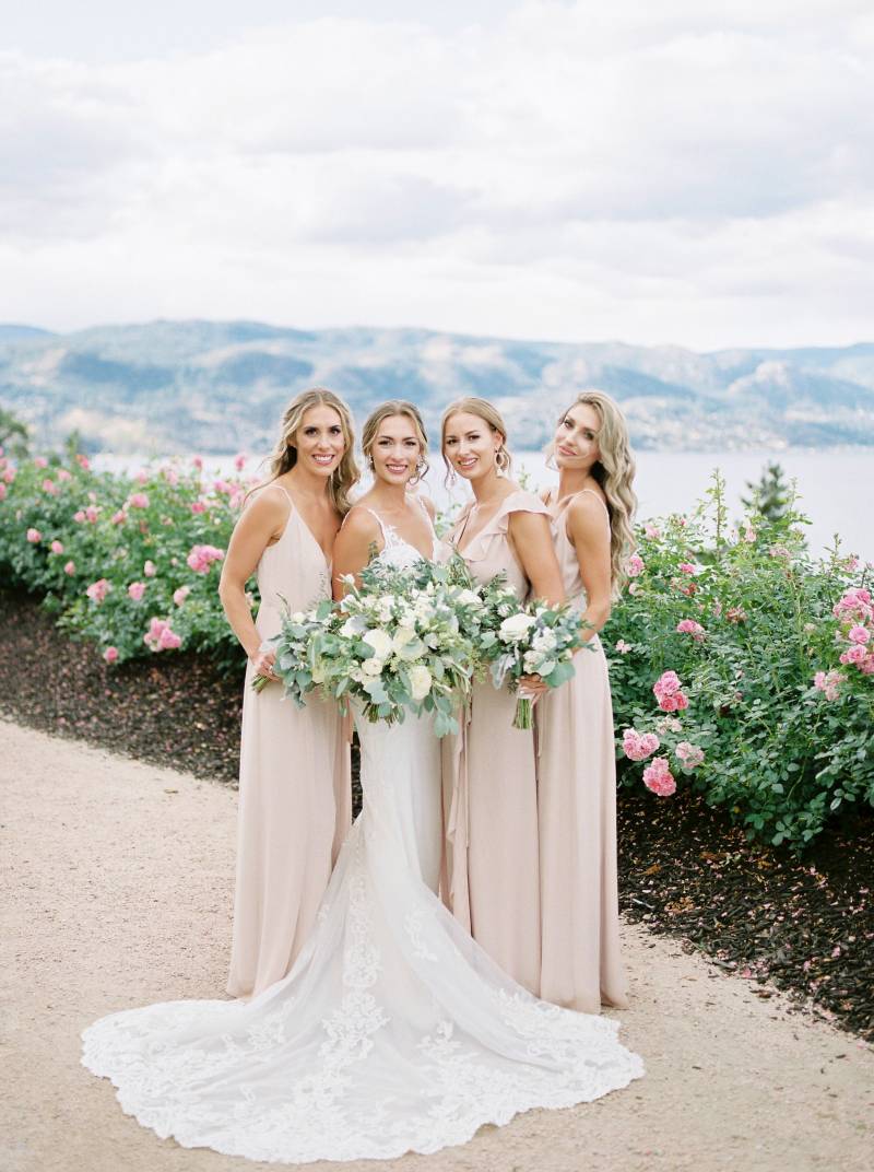 Bride standing with bridesmaids holding white flower bouquets on dirt path in front of pink rose bushes 