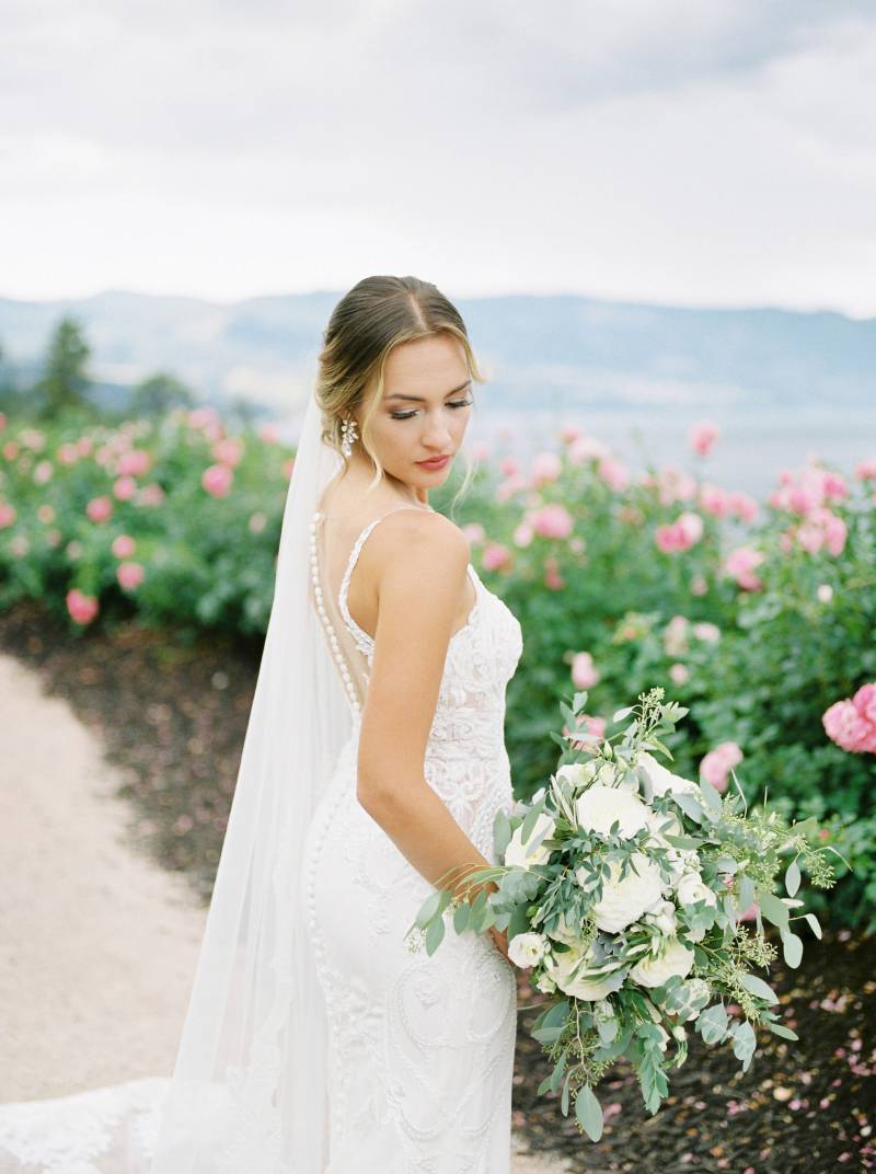 Bride looking over shoulder holding white bouquet in front of pink rose bushes 