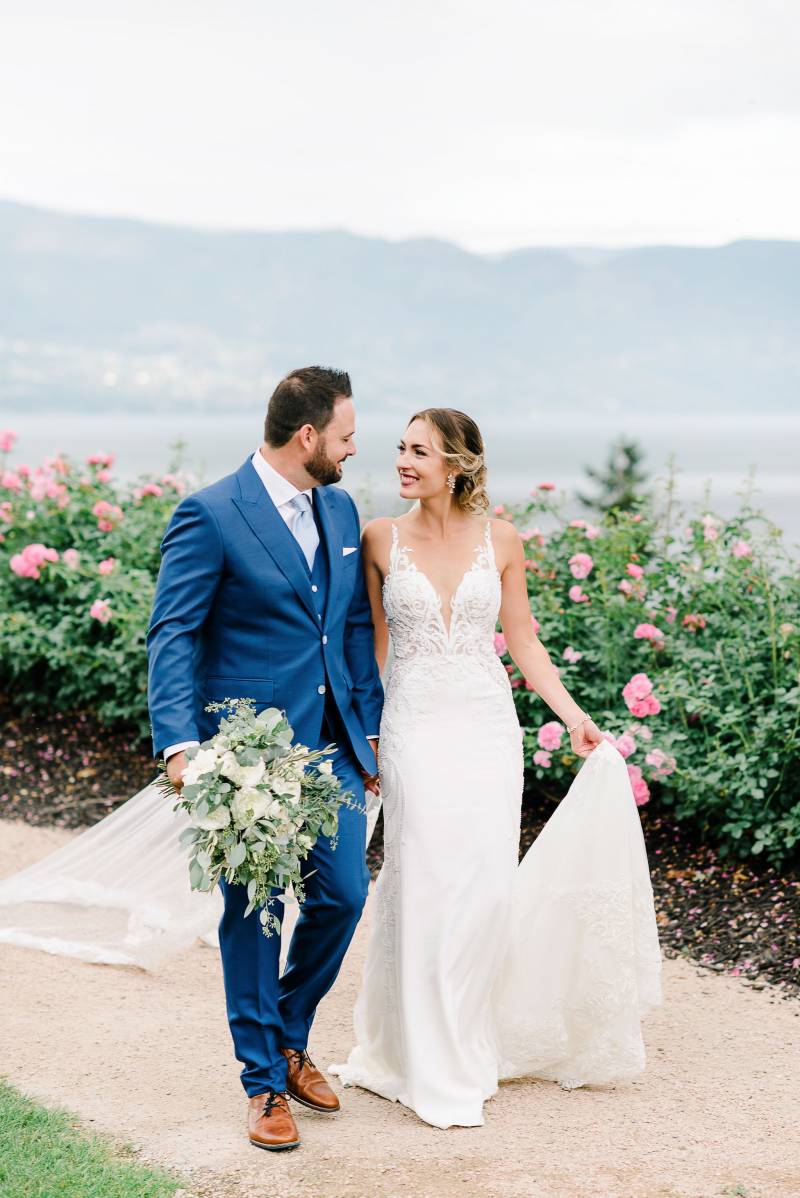 Bride and groom walk together down dirt path beside pink rose bushes 