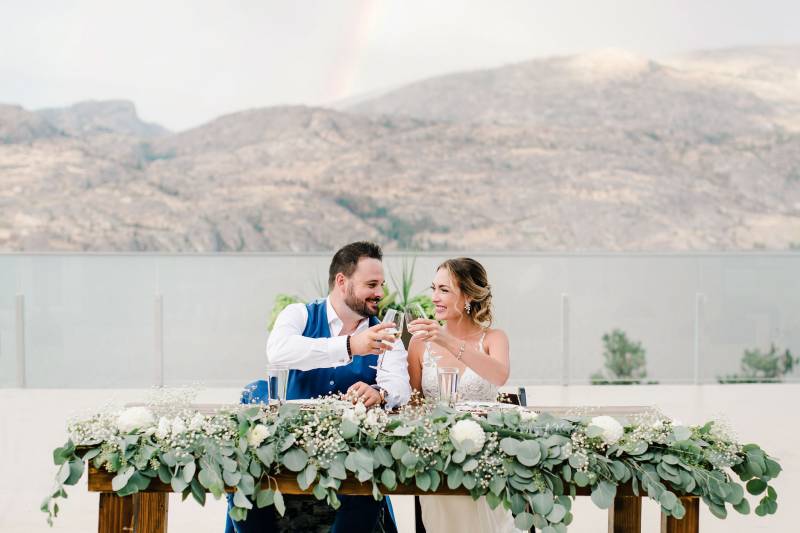 Bride and groom cheers sitting at head table with green and white arrangement on terrace overlooking mountains