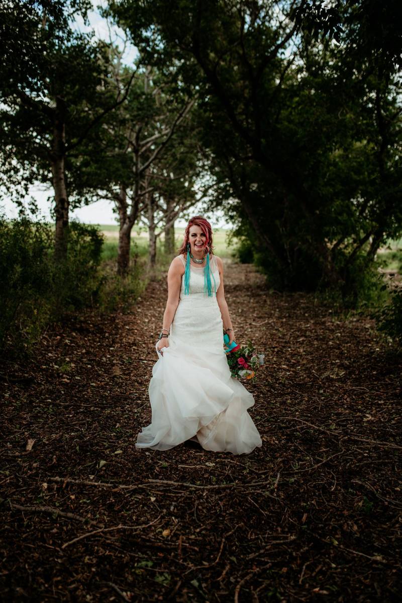 Bride walks in white dress holding multicolored bouquet down forest pathway  