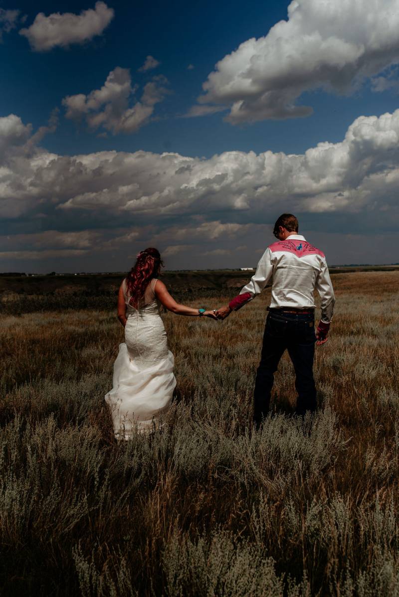 Bride and groom walk holding hands in large open grassy field 