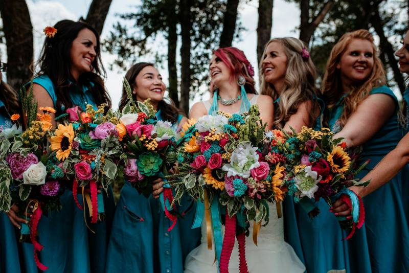 Bride and bridesmaids hold out teal  yellow and pink bouquets smiling in line 