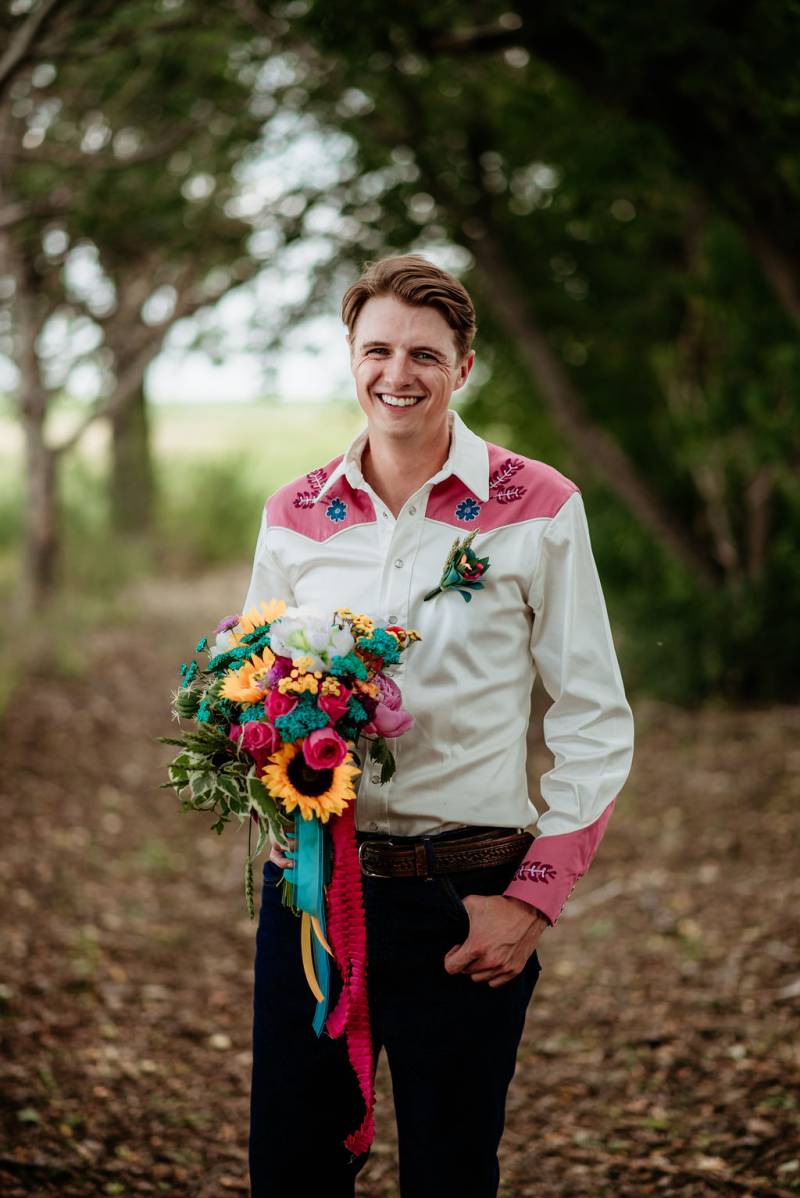 Groom holding bouquet wearing pink and white embroidered dress shirt stands smiling on forest pathway 