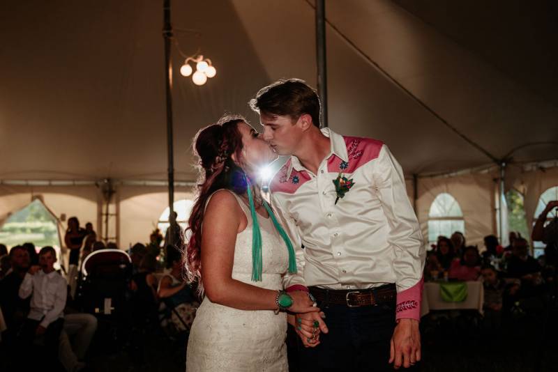 Bride and groom kiss under white tent while guests sit in background 