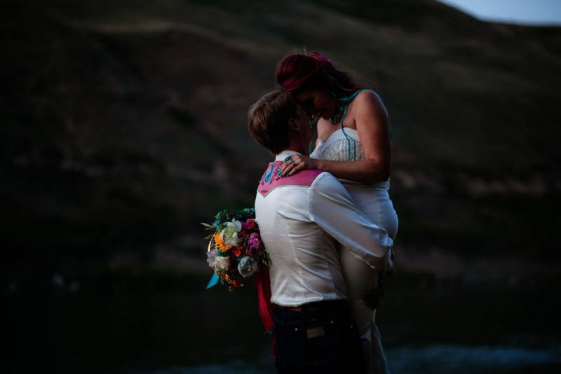 Groom lifts bride touching foreheads holding multicolored bouquet at dusk 