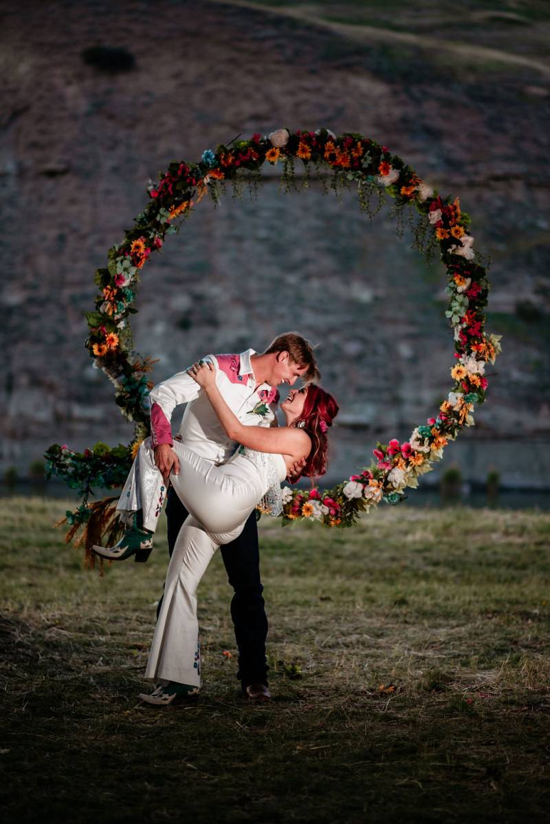 Groom dips bride smiling in front of yellow crimson and teal floating wedding arch 