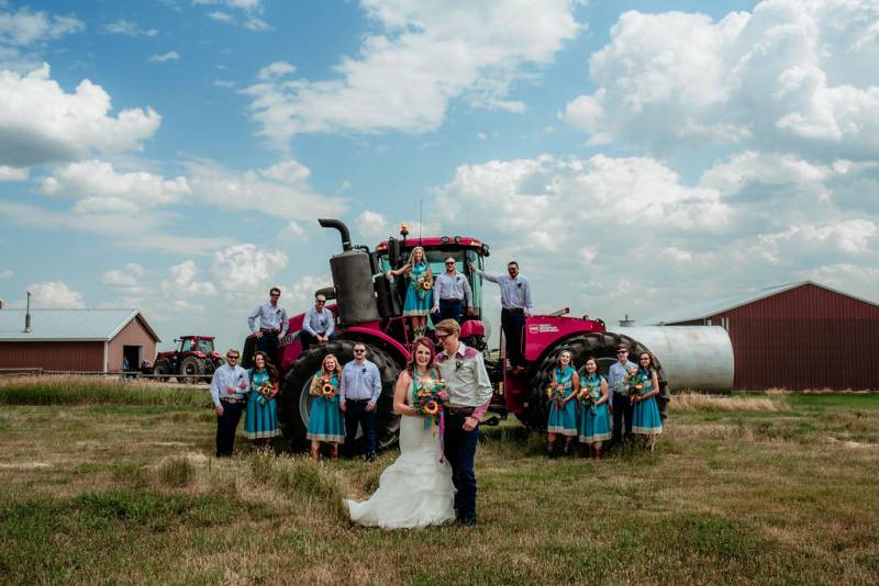Bride and groom stand together holding bouquet in front of bridesmaids and groomsmen on and around red tractor 