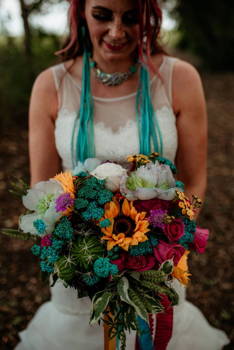 Bride in white dress holds teal yellow crimson and white bouquet 