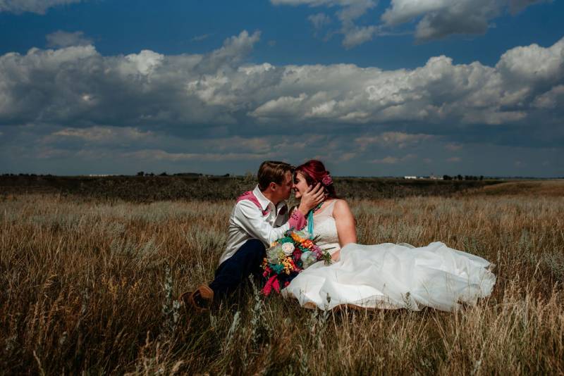 Bride and groom sit in grassy field  groom touches cheek 