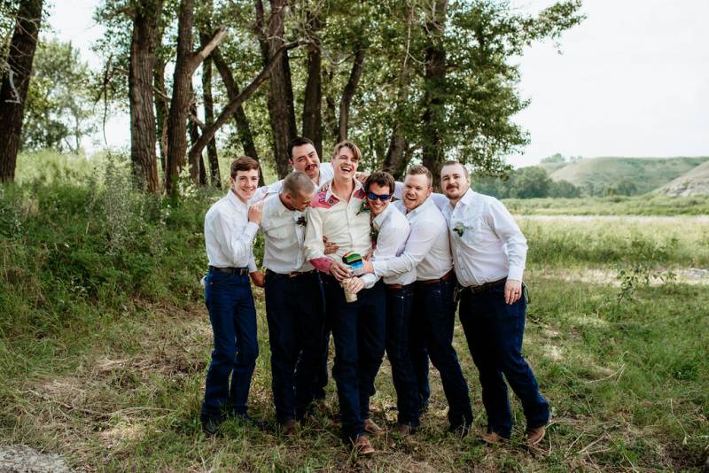 Groom stands in between groomsmen arms over shoulders smiling and laughing in grassy field 