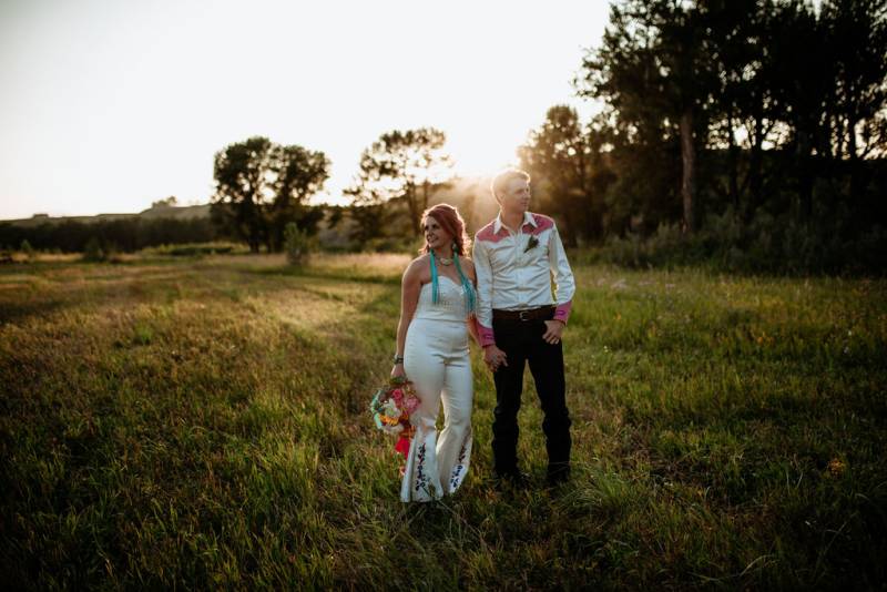 Bride and groom stand holding hands in grassy field at sunset 